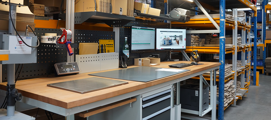 Modern eingerichteter Packarbeitsplatz in einem Lager mit Holztischplatte, zwei Monitoren, Verpackungsstation, Waage, Kartonagen und Regalsystemen im Hintergrund.