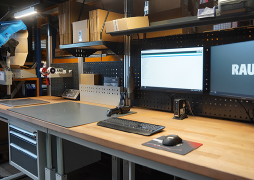 Arbeitsplatz im Warenausgang mit RAU Werkbank, Holzplatte, zwei Monitoren und Ablagefächern.