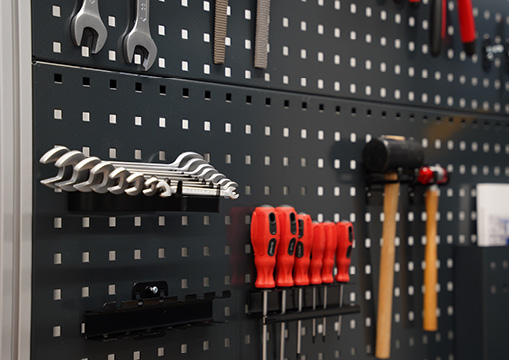 Dunkle Lochwand mit aufgehängtem Werkzeug – darunter Ringschlüssel-Set, Schraubendreher mit roten Griffen und ein Hammer – als Werkbank-Zubehör in einer Werkstatt.