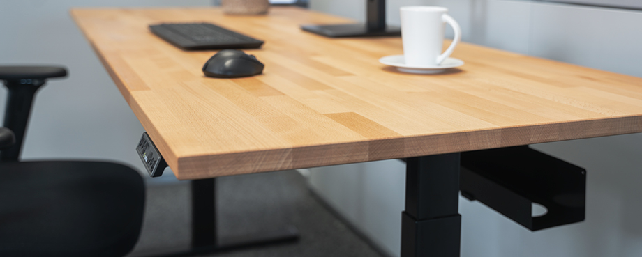Detailaufnahme eines höhenverstellbaren Schreibtischs mit Buche-Massivholzplatte, Tastatur, Maus und Kaffeetasse – ergonomischer Arbeitsplatz.