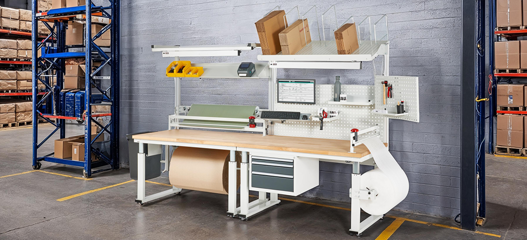 Höhenverstellbarer Verpackungsarbeitsplatz mit Buche-Arbeitsplatte, Lochplatte, Abrolleinheit, Schneidgerät, Kartonagenmagazin, LED-Arbeitsplatzleuchte und Schubladenschränken – ideal für effiziente Verpackungsprozesse.
